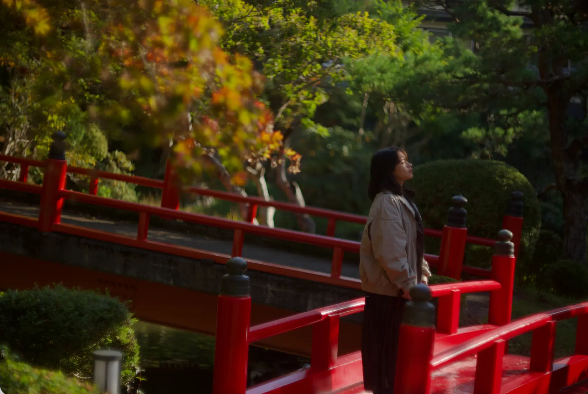 A woman standing on a red bridge in a sunlit Japanese garden